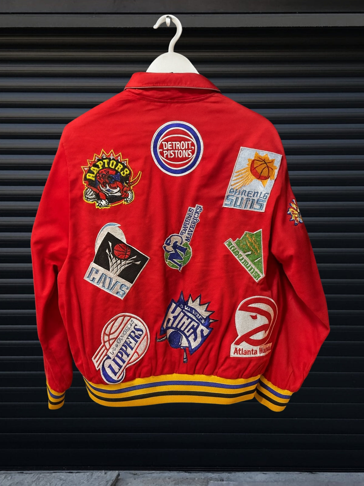 Vintage Red NBA patch jacket with shirt collar and red striped hem featuring team logos including Lakers, Clippers, Bucks, Suns, 76ers, Magic and more, hanging on hanger