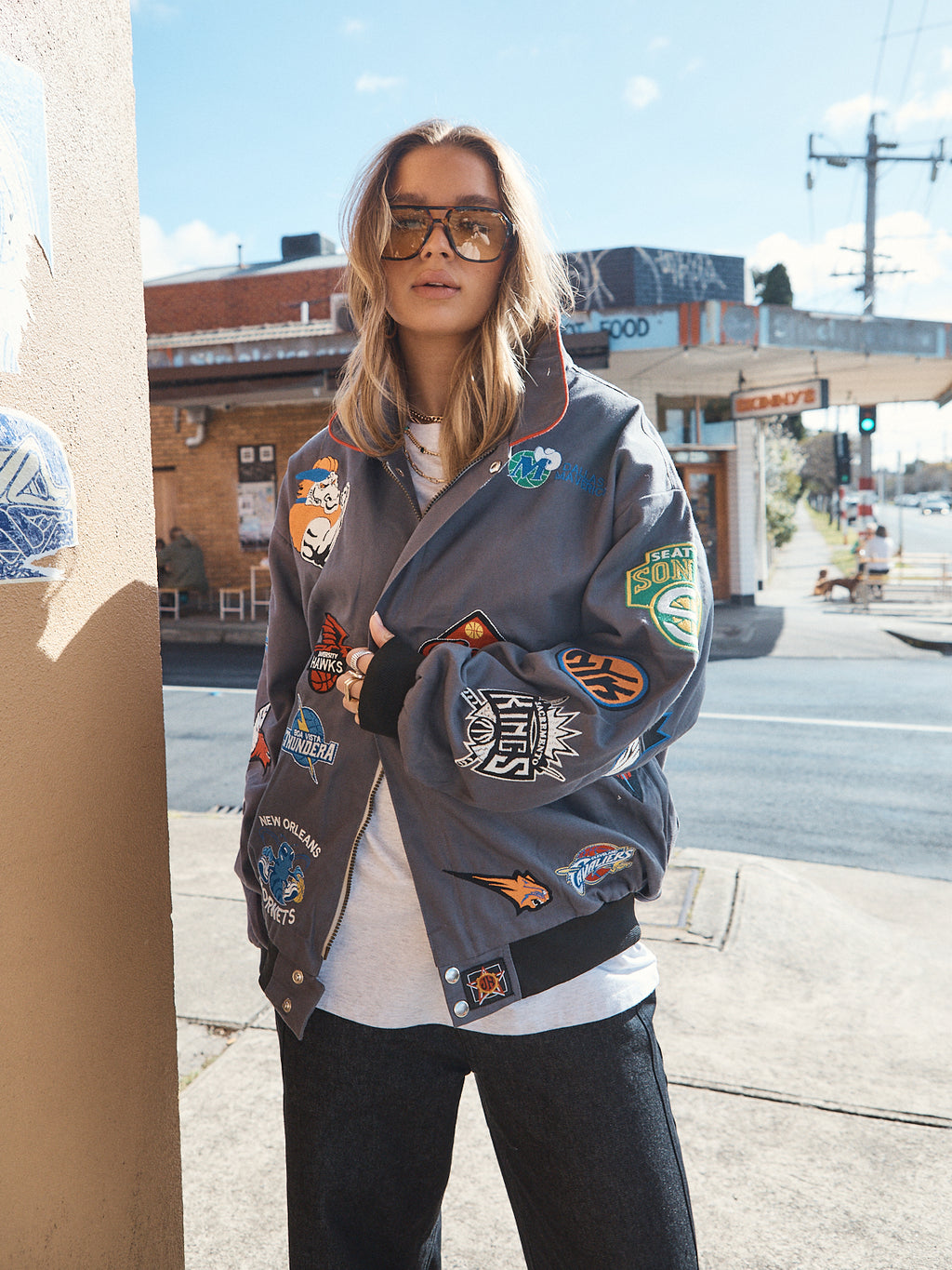 Woman wearing oversized grey NBA patch jacket layered over unisex grey marle t-shirt and wide-leg jeans, streetwear style