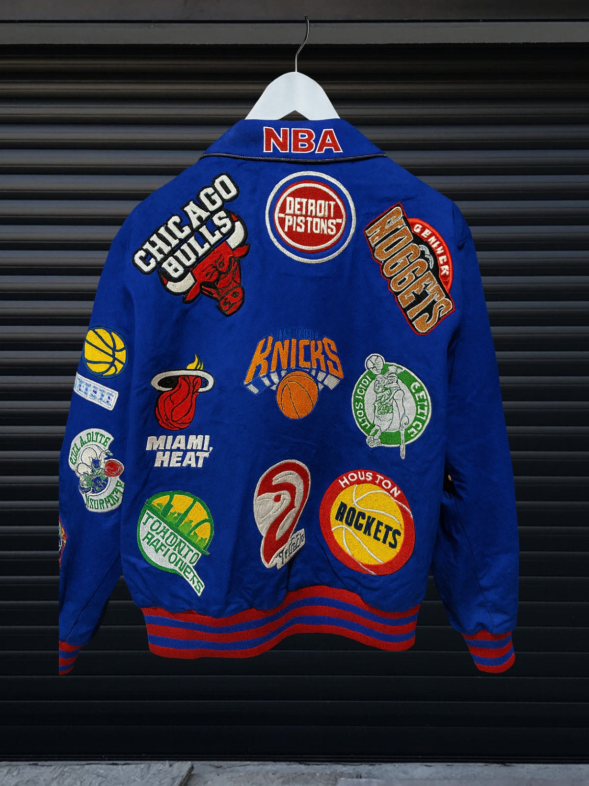 Vintage blue NBA patch jacket with shirt collar and red striped hem featuring team logos including Lakers, Clippers, Bucks, Suns, 76ers, Magic and more, hanging on hanger
