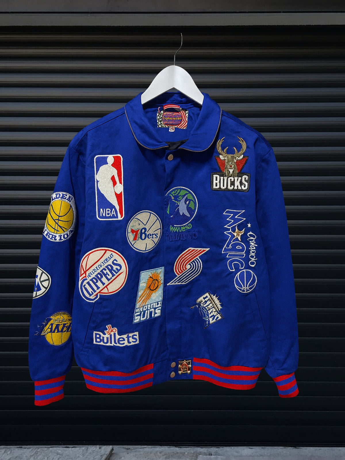 Vintage blue NBA patch jacket with shirt collar and red striped hem featuring team logos including Lakers, Clippers, Bucks, Suns, 76ers, Magic and more, hanging on hanger