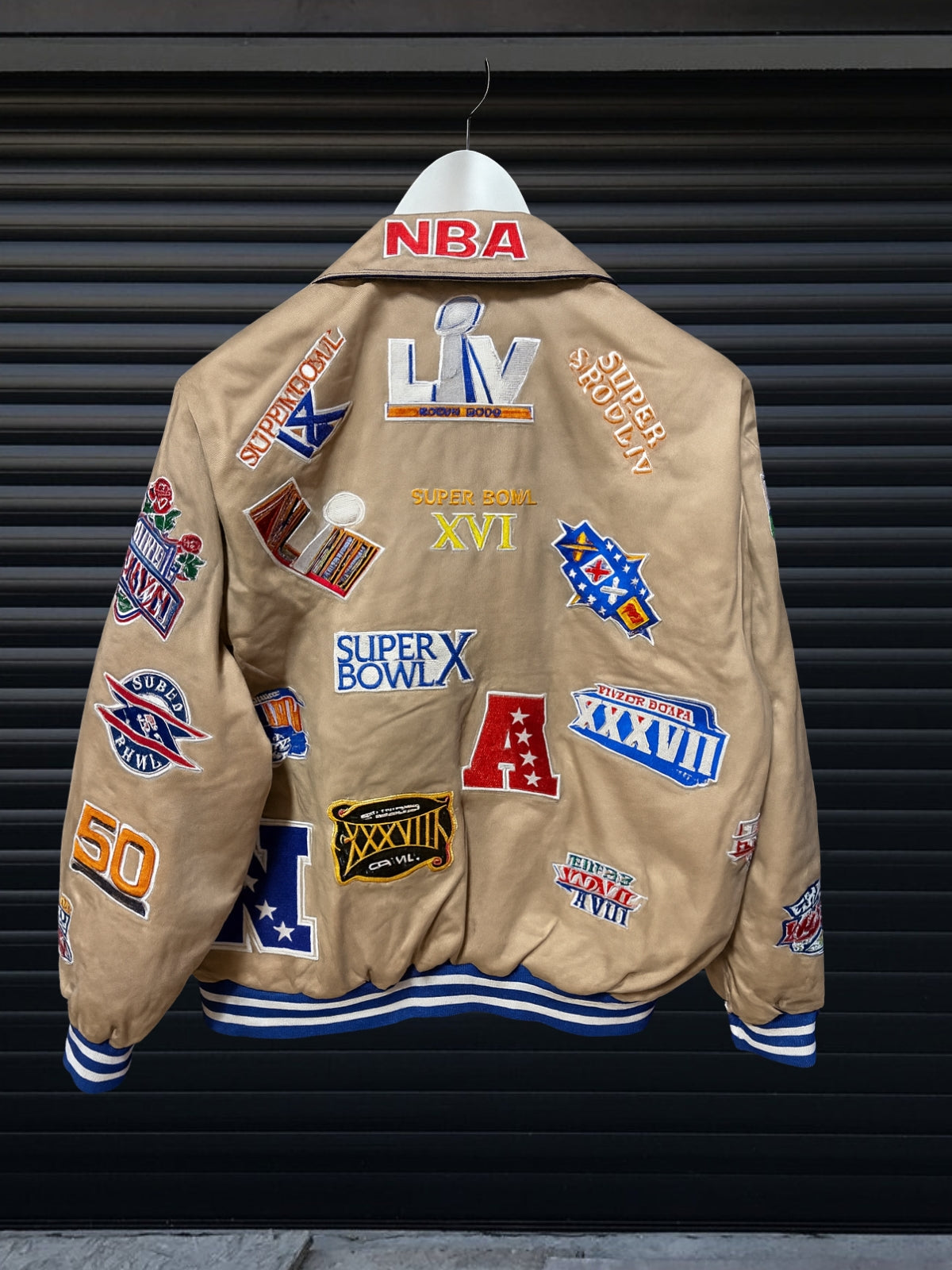 Vintage khaki NBA patch jacket with shirt collar and red striped hem featuring team logos including Lakers, Clippers, Bucks, Suns, 76ers, Magic and more, hanging on hanger